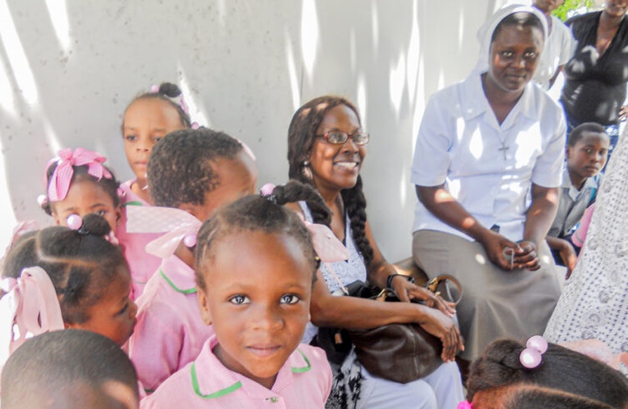 Young girls in pink school uniforms sit with smiling women from their community in the shade
