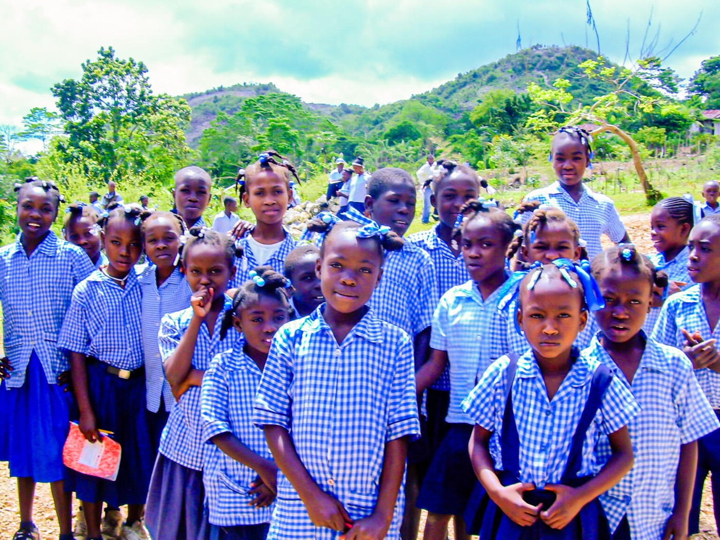 A large group of Haitian schoolchildren in blue gingham uniforms gathered outdoors beneath green hills