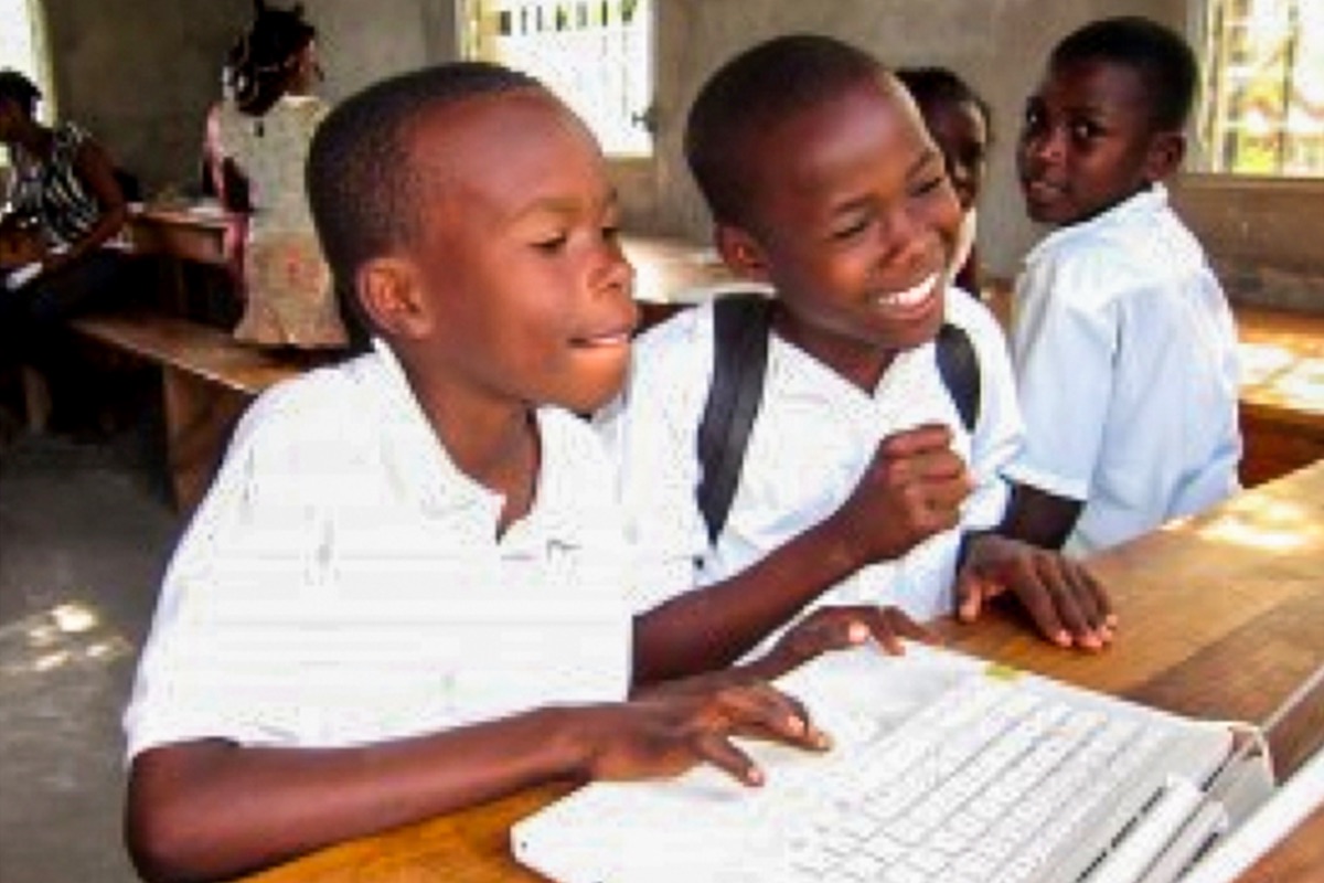 Two laughing schoolboys in white uniforms share a laptop at a wooden classroom desk while classmates look on